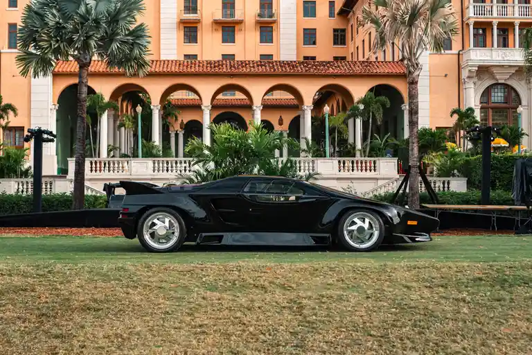 A black, angular, futuristic sports car with a large rear wing and silver wheels is parked on a grassy lawn in front of a…