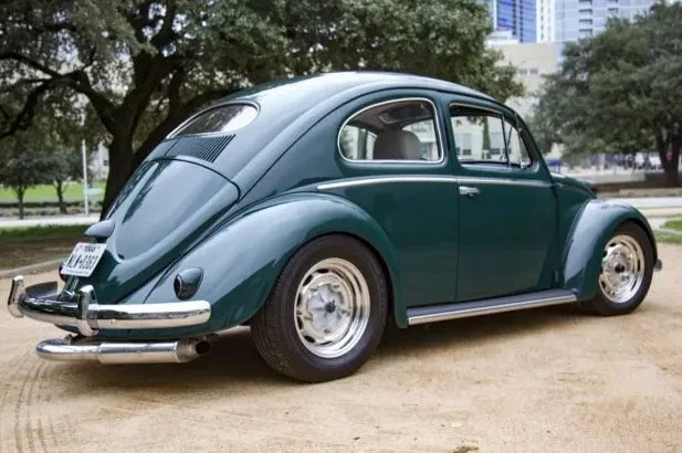 A dark green classic Volkswagen Beetle is parked on dirt with trees and buildings in the background. The car has chrome…
