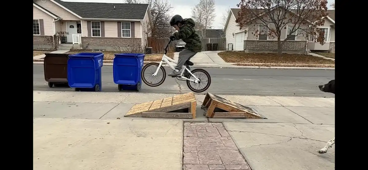 A young person wearing a helmet and camouflage jacket rides a white BMX bike mid-air over two homemade wooden ramps on a…