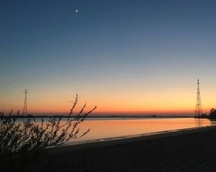 A serene sunset over a calm body of water with silhouetted power lines and a crescent moon visible in the sky. The…