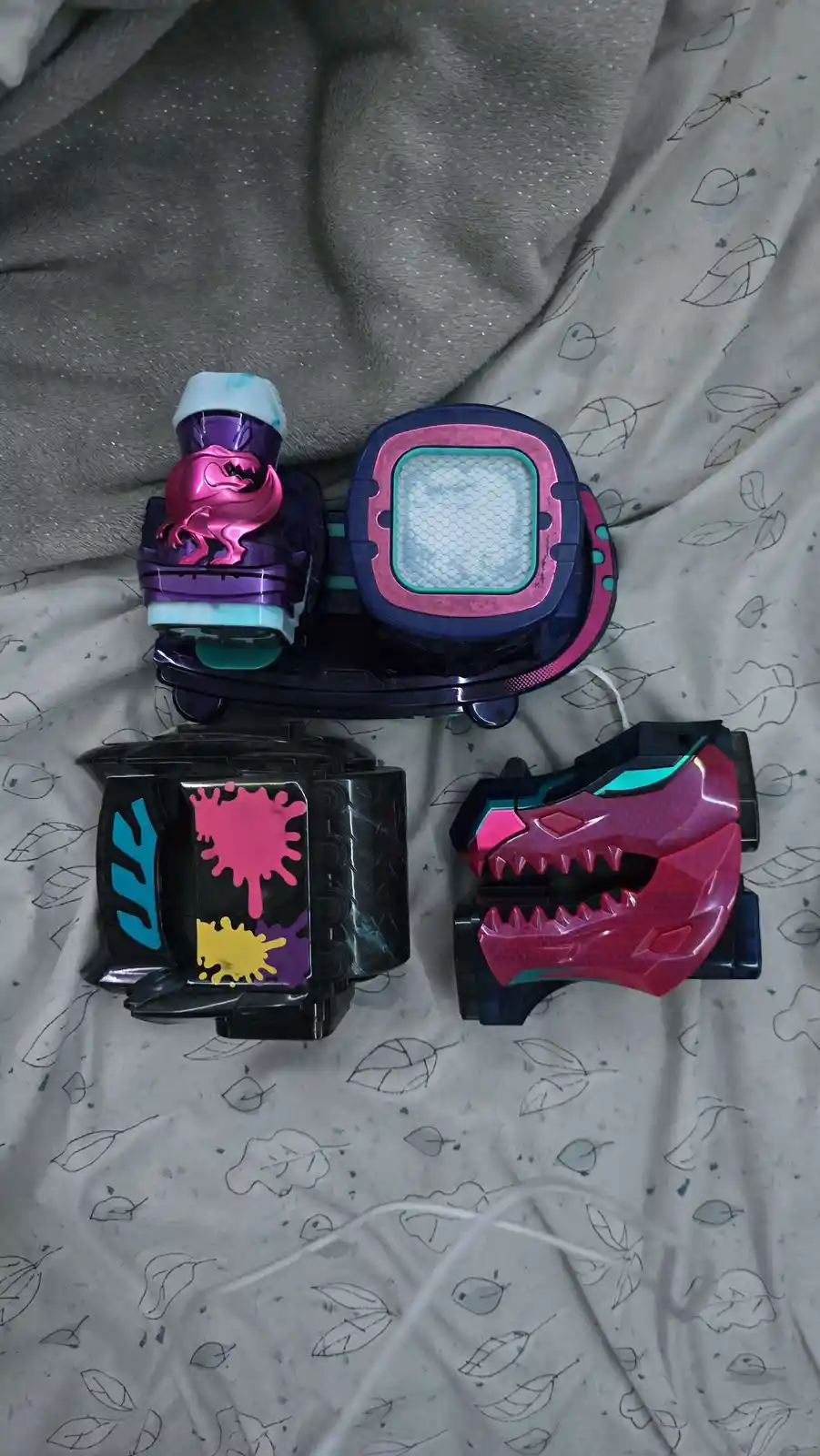 Four colorful Kamen Rider toy transformation devices are laid out on a grey and white leaf-patterned fabric. The top device…
