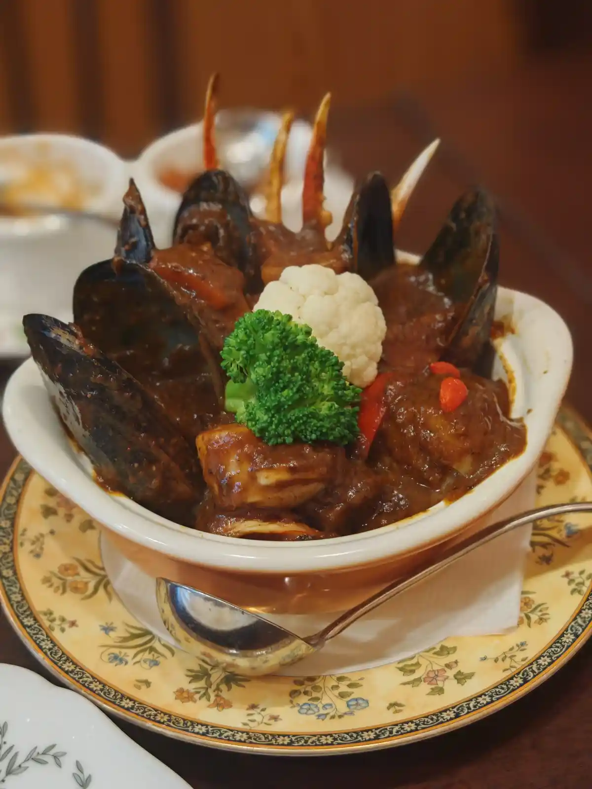 A close-up photo shows a white ceramic bowl filled with mussels, crab claws, and chunks of meat in a dark, thick sauce. The…