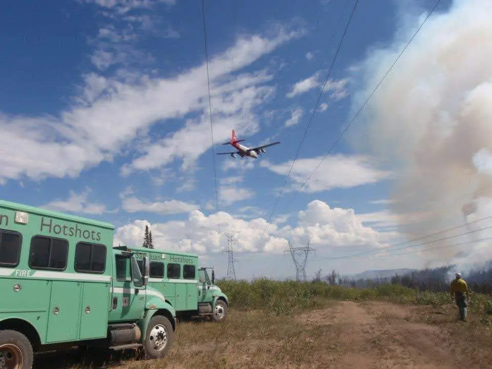 Two green fire department buses labeled 'Hotshots' are parked on a dirt road near a wildfire with smoke rising in the…