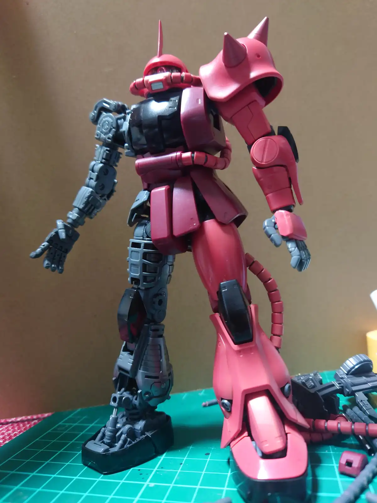 A red and grey robot model, resembling a Gundam, stands on a green cutting mat with its left leg missing and replaced by a…