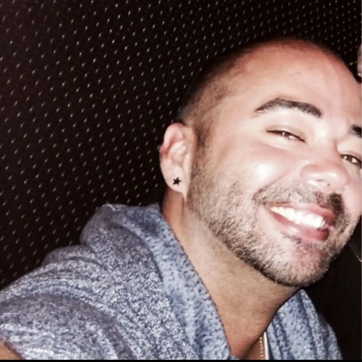 a close-up photo of a smiling man with a short buzz cut and stubble, wearing a grey hoodie, seated against a dark perforated…