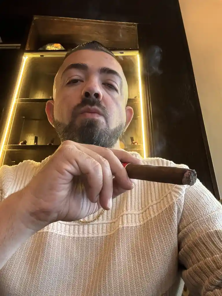A man with a beard and short hair, wearing a light-colored ribbed sweater, holds a lit cigar in his right hand and looks…