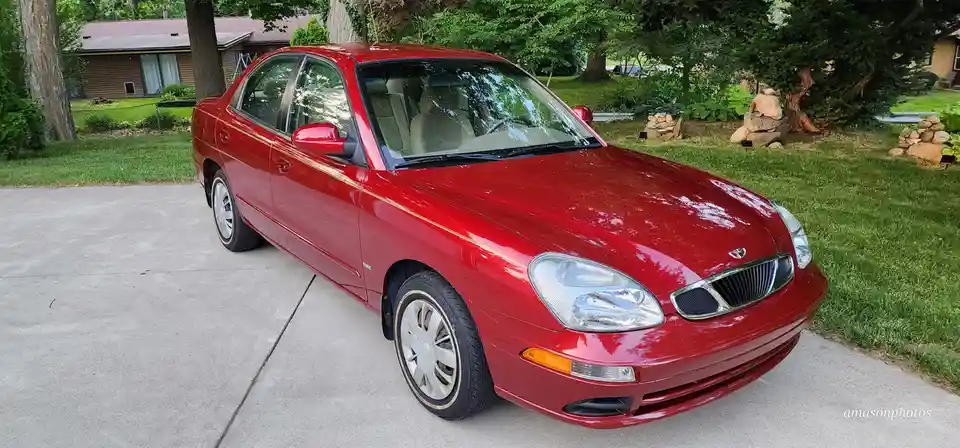 A glossy red 2000s-era sedan, likely a Kia Spectra or similar model, is parked on a concrete driveway in a suburban yard…