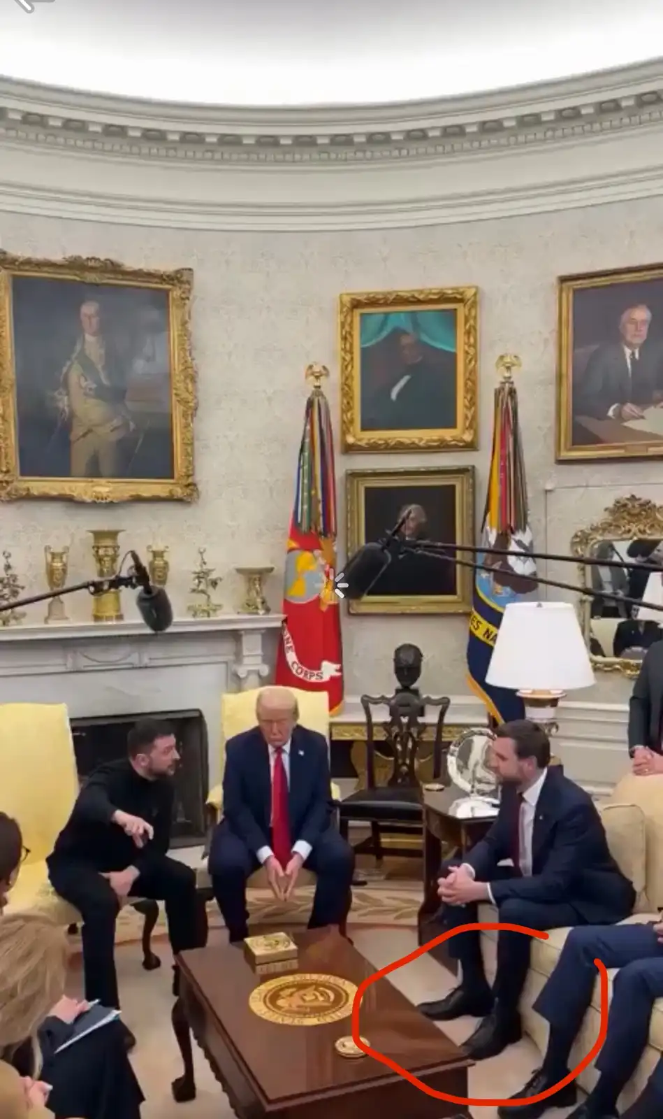 In the Oval Office, surrounded by presidential portraits and flags, two men sit on a couch with their feet visible under the…