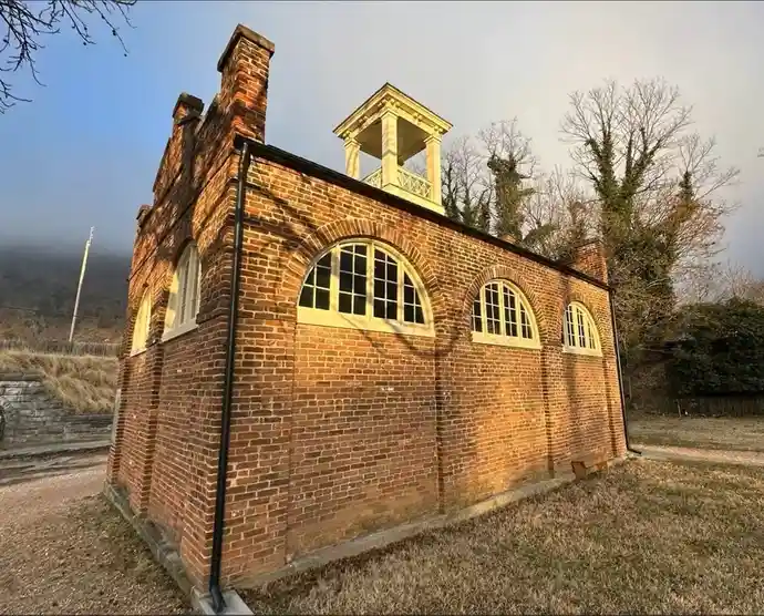 a small brick building with arched windows and a cupola on top, set in a grassy area with trees in the background, overlaid…