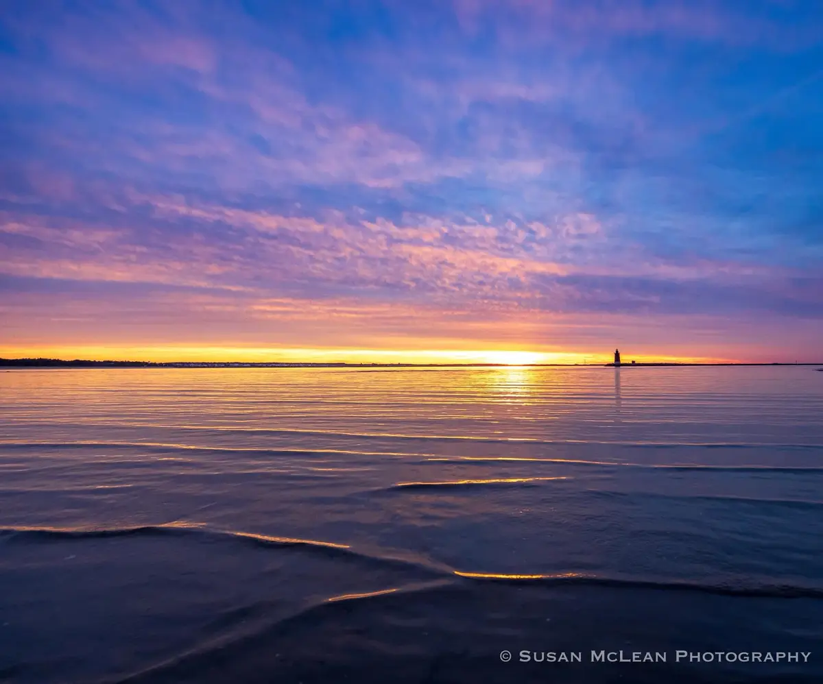 A serene sunset over water with a lighthouse in the distance, featuring a sky painted with purple, pink, and orange hues…