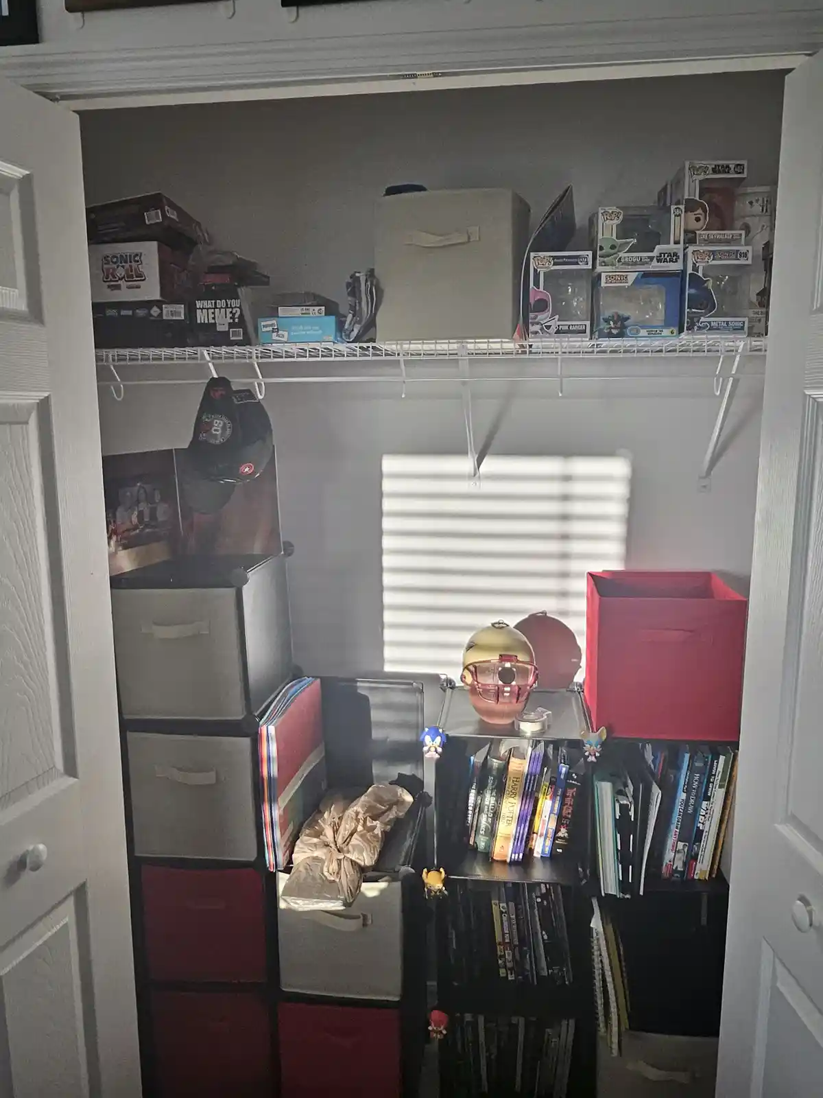 A cluttered closet filled with storage bins, Funko Pops, books, and collectibles, with a box labeled 'WHAT DO YOU MEME?'…