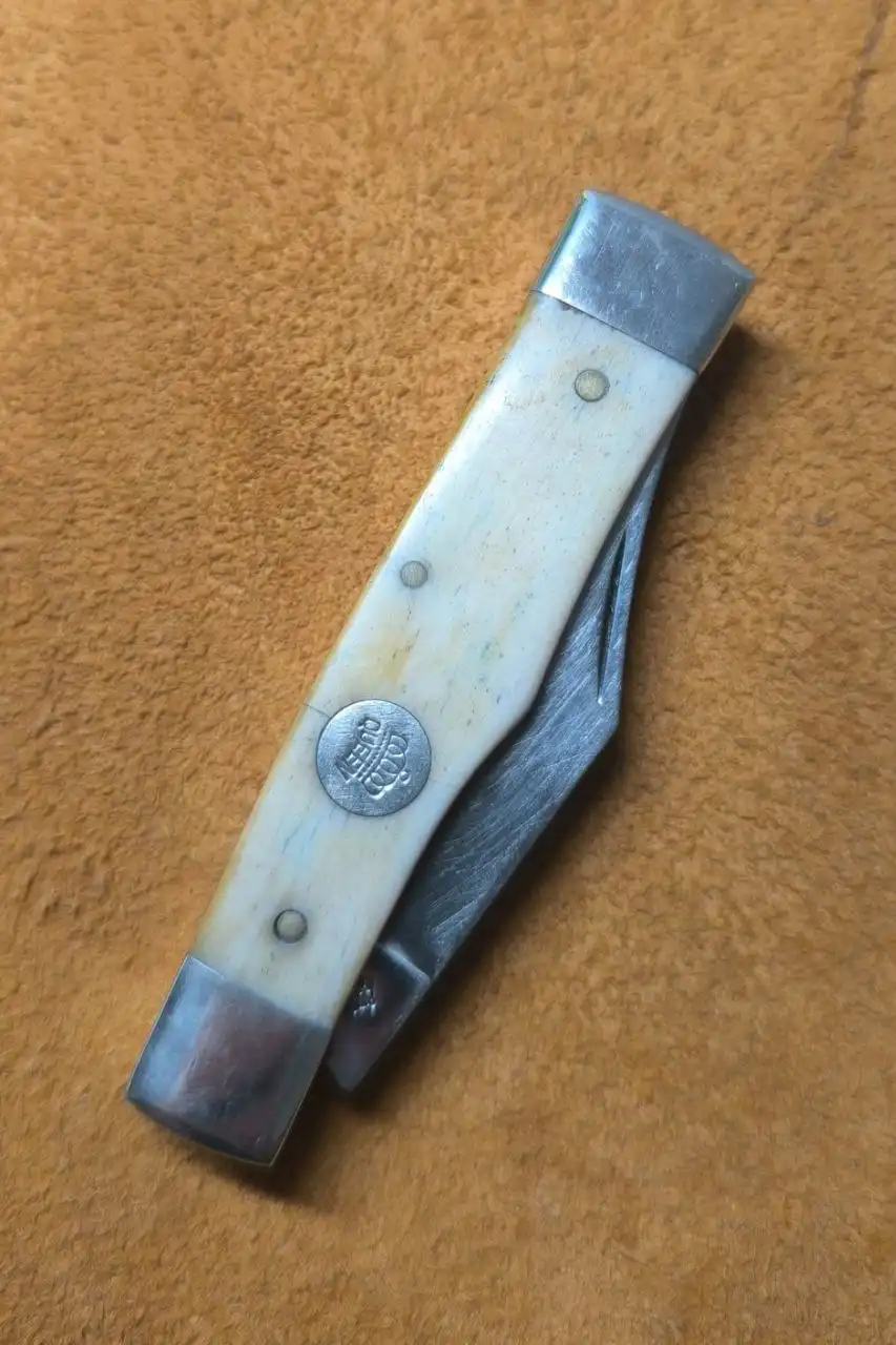a close-up photo of a folding knife with a bone or ivory handle and metal bolster, resting on a textured brown leather…