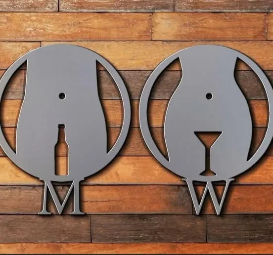 Two gray metal cutouts shaped like dog heads hang on a wooden wall; the left one has a bottle-shaped cutout for a mouth and…