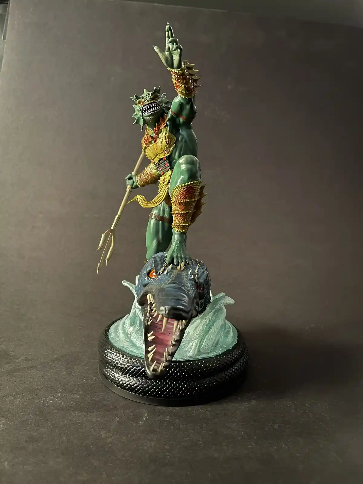 A green-skinned, reptilian sea god statue stands triumphantly on the head of a large, open-mouthed crocodile-like creature…