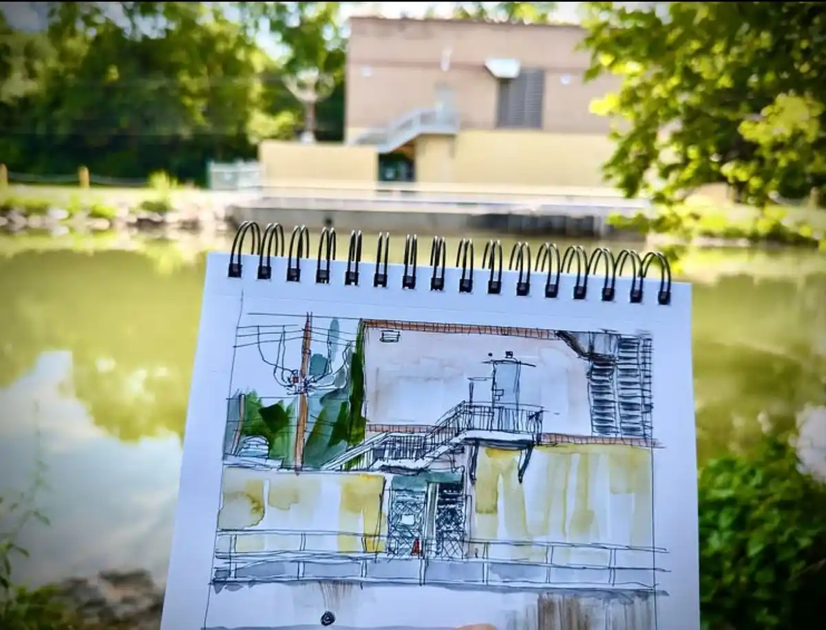 A spiral-bound sketchbook is held up outdoors, showing a watercolor and ink drawing of a building with stairs and railings…