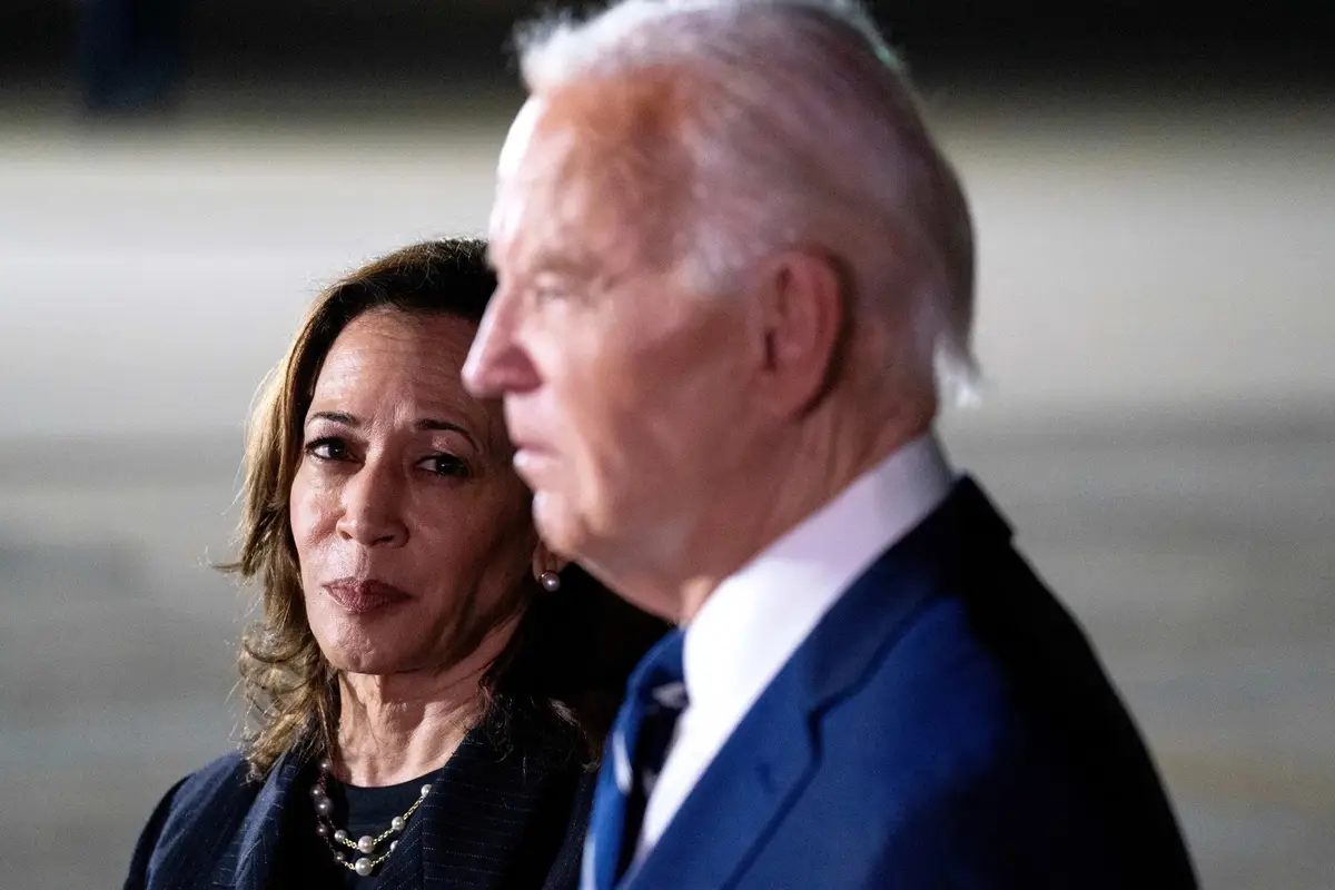 A photo shows Kamala Harris looking intently forward while standing beside Joe Biden, who is in profile. Harris wears a dark…