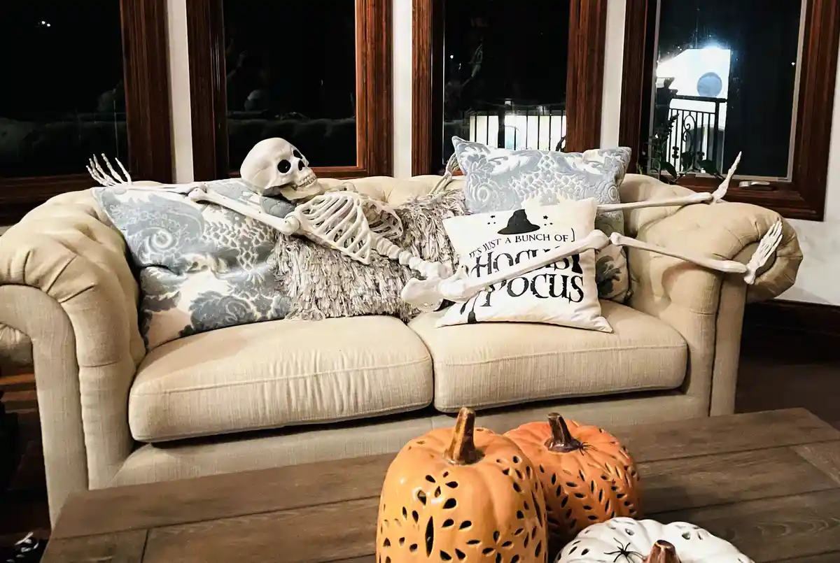 A white plastic skeleton is lounging on a beige couch, propped up by patterned pillows, with its arms draped over the…