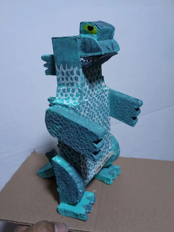 A handmade, teal-colored papier-mâché monster sculpture with a blocky, geometric design, sharp teeth, and a single visible…