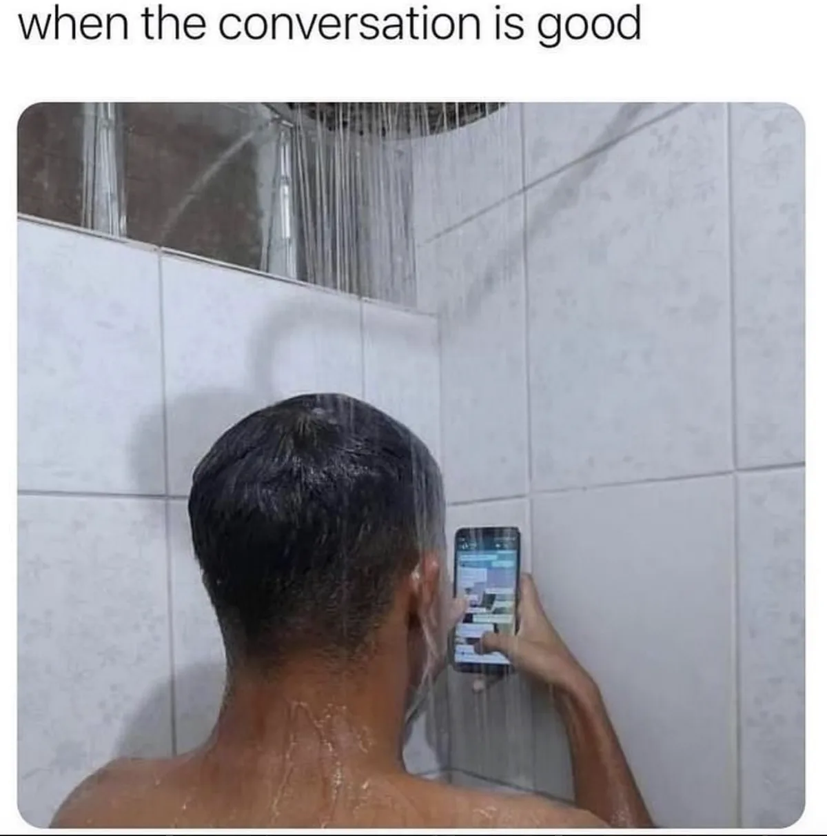 A person is seen from behind, standing in a shower with water falling on their head and back, holding a smartphone in their…