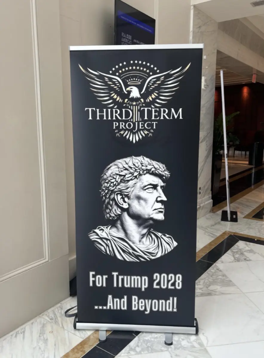 A black banner stand in a marble lobby displays a logo of an eagle with stars above the text 'THIRD TERM PROJECT'. Below, a…