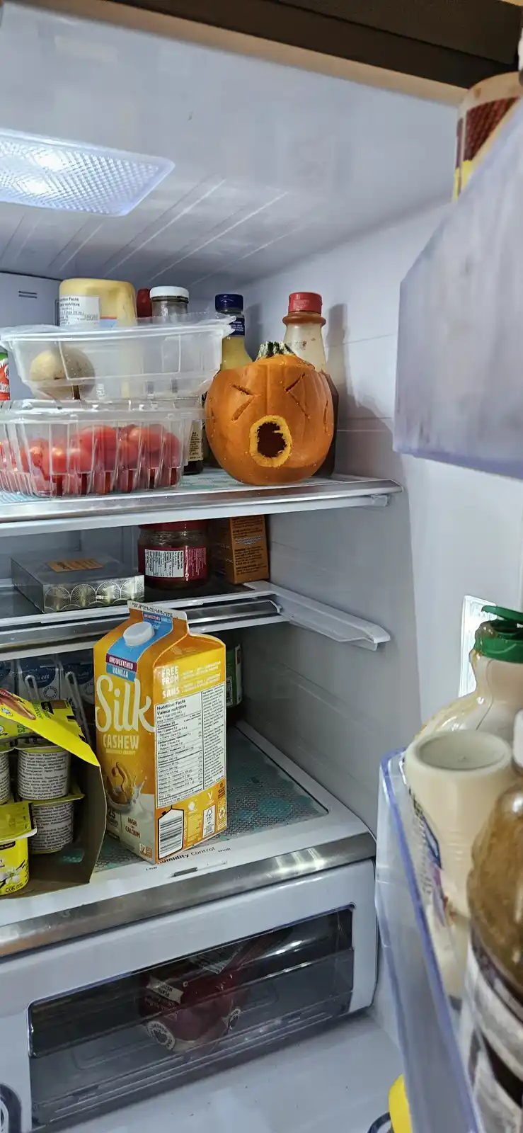 A refrigerator interior shows a carved pumpkin with a distressed face on a top shelf, surrounded by various food items like…