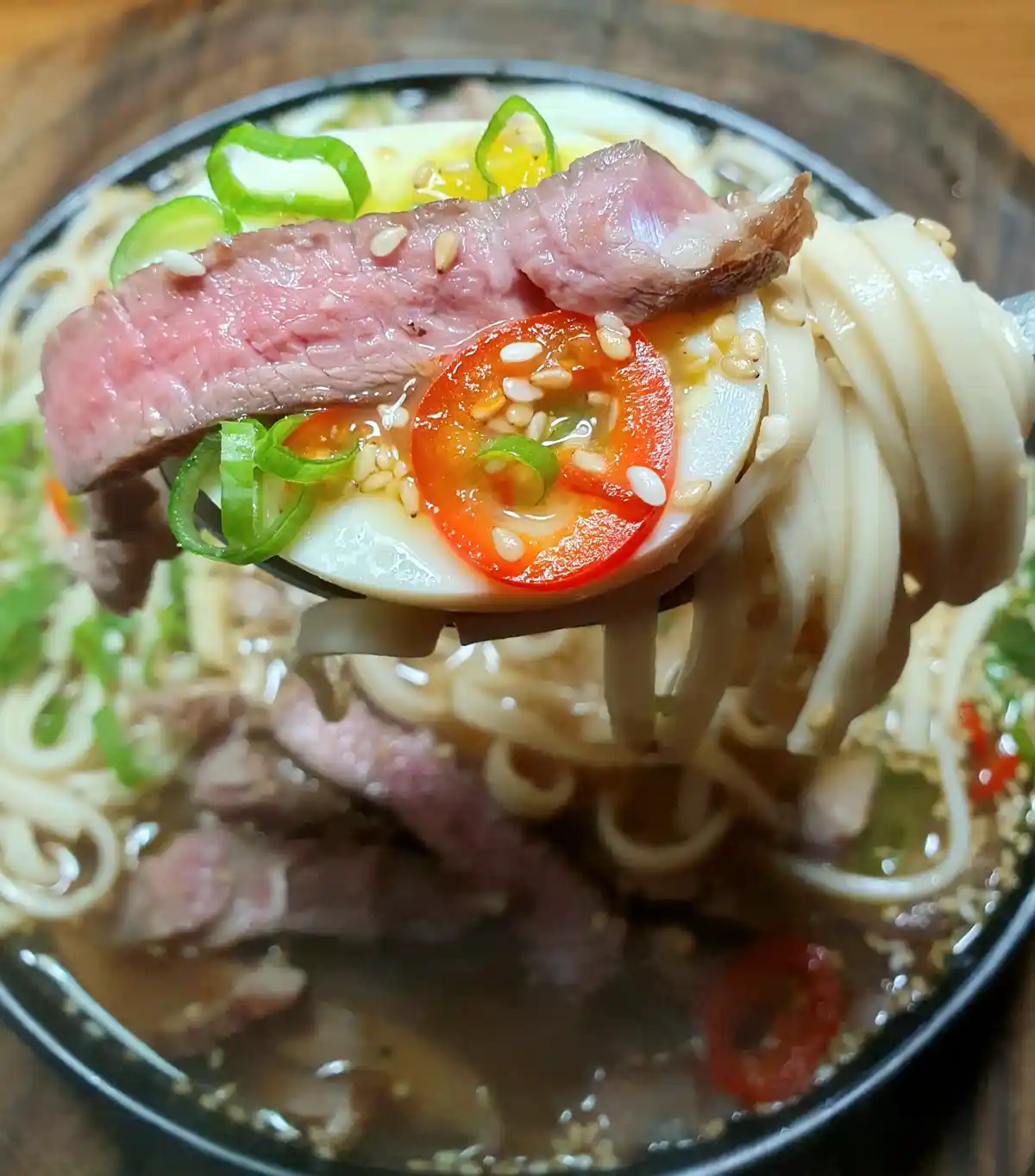 a close-up photo shows a fork lifting a mouthful of ramen from a black bowl, featuring thick noodles, a slice of medium-rare…