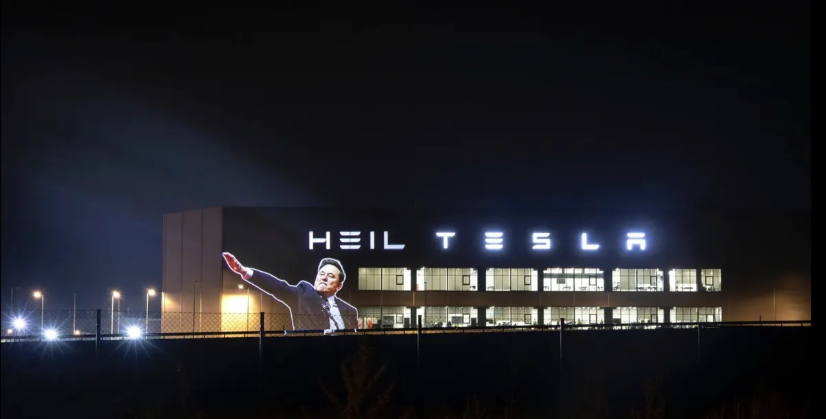 A nighttime photo of a large building with illuminated windows and a sign that reads 'HEIL TESLA'. Projected onto the side…