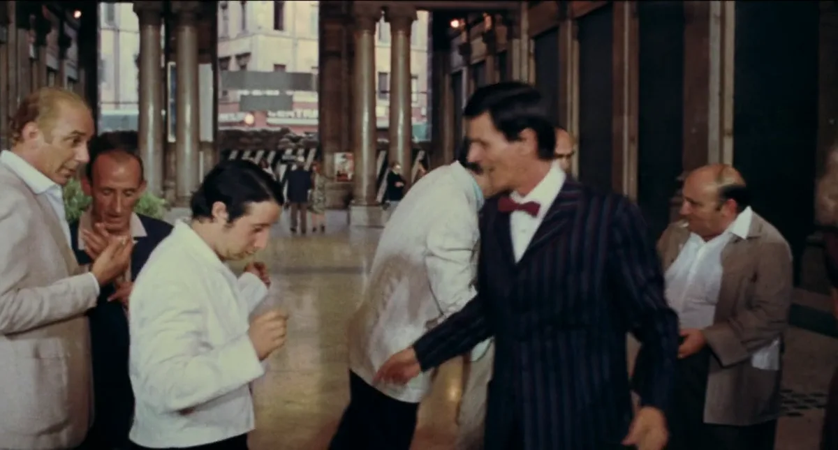A still from a 1970s film shows a man in a pinstripe suit and red bow tie smiling and gesturing toward a younger man in a…