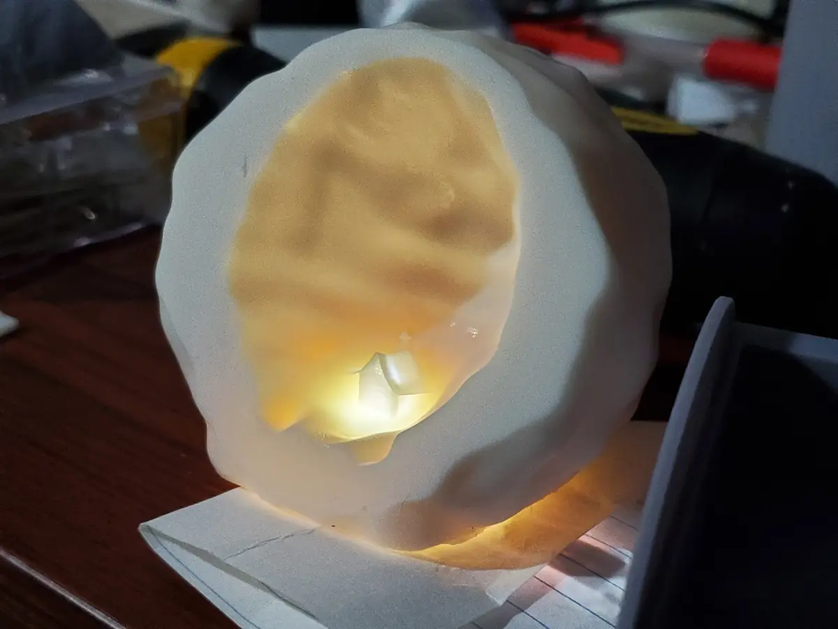 A 3d printed object shaped like a fried egg sits on a desk, glowing from within. The translucent white shell encases a…