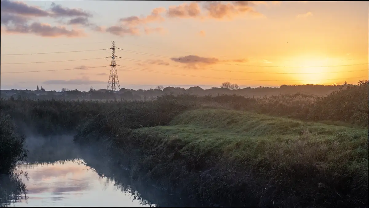 A serene landscape photo of a misty river at sunrise, with orange and pink clouds reflecting on the water and a power line…