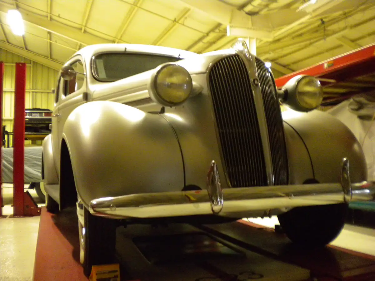 A silver classic car with a prominent grille and round headlights is shown from the front, elevated on a hydraulic lift…