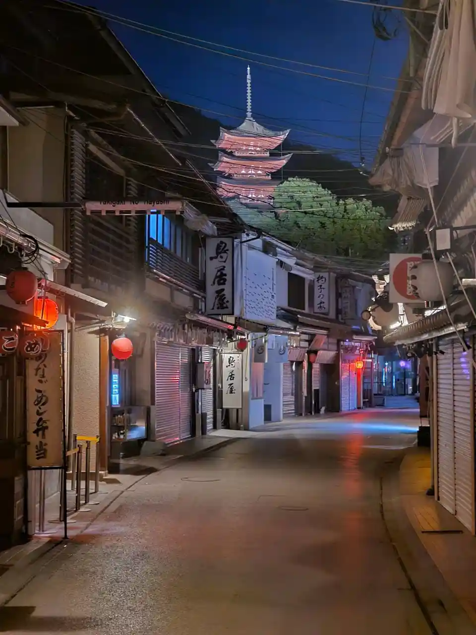 A nighttime photo of a quiet, narrow Japanese street with closed shop shutters, red lanterns, and a distant pagoda visible…