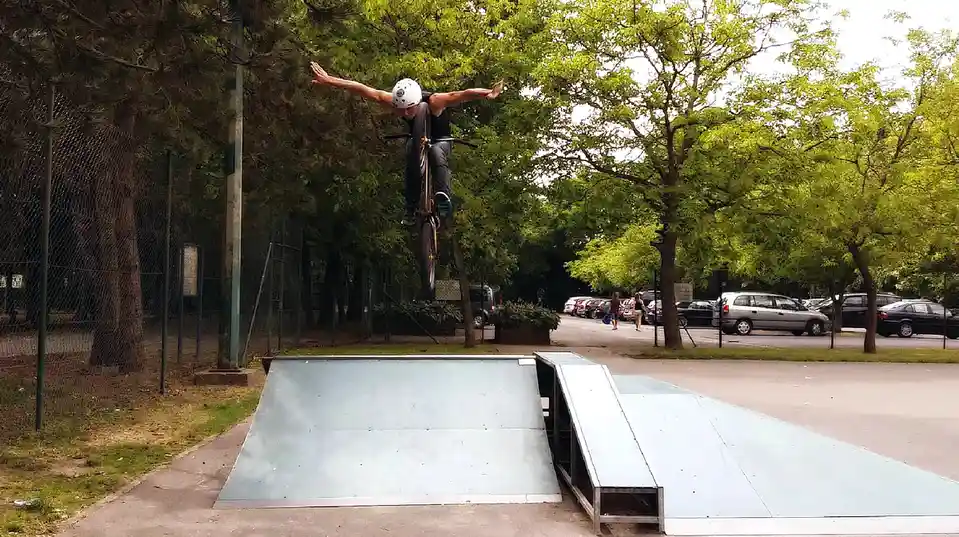A person wearing a white helmet and dark clothing is airborne on a BMX bike above a metal ramp in a park, arms spread wide…