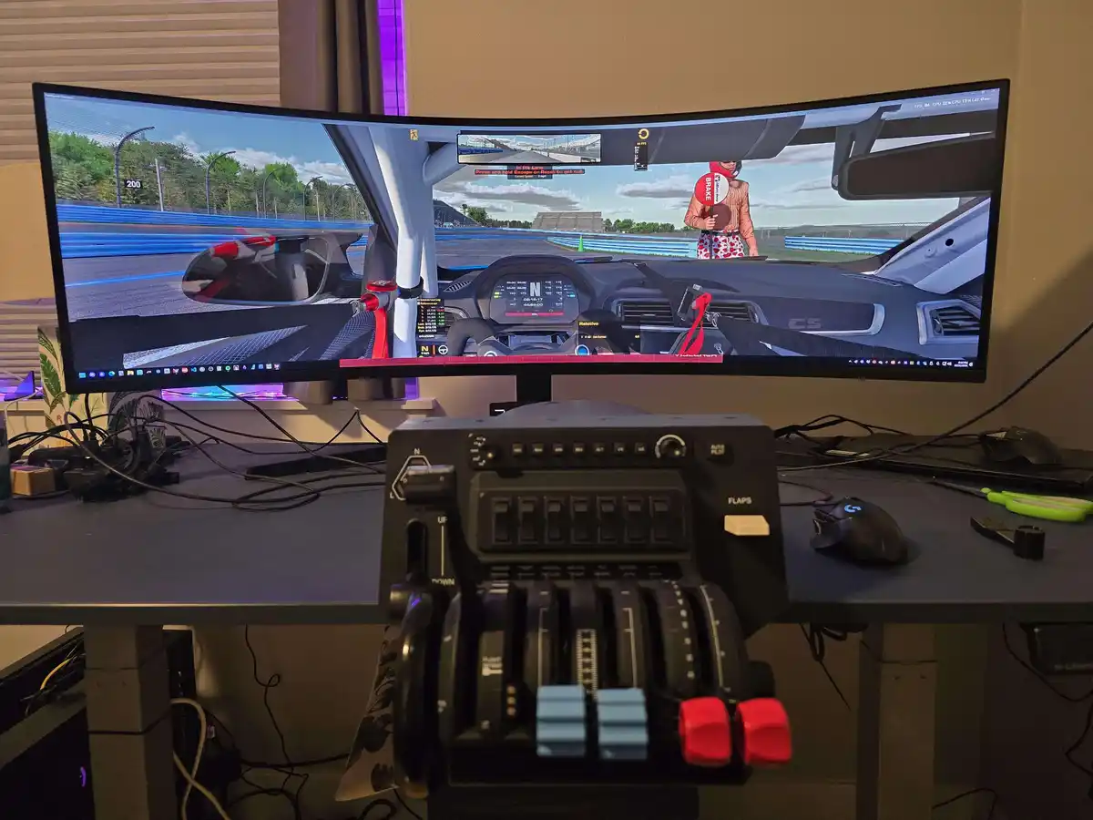 A curved monitor displays a racing game from a driver's perspective, showing a pit lane with a person holding a 'BRAKE'…