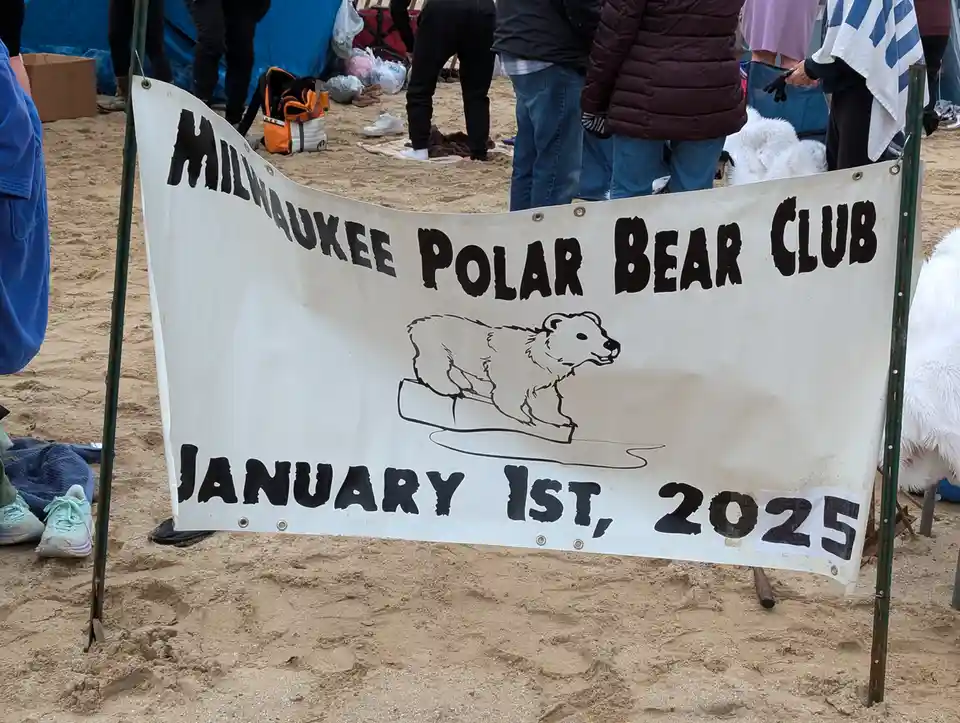 A white banner on a sandy beach reads 'MILWAUKEE POLAR BEAR CLUB' with a line drawing of a polar bear on ice, and 'JANUARY…