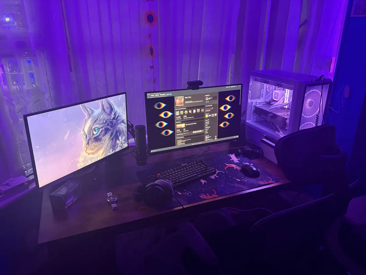 A dimly lit gaming setup bathed in purple light features two monitors: one displays a detailed illustration of a cat, while…