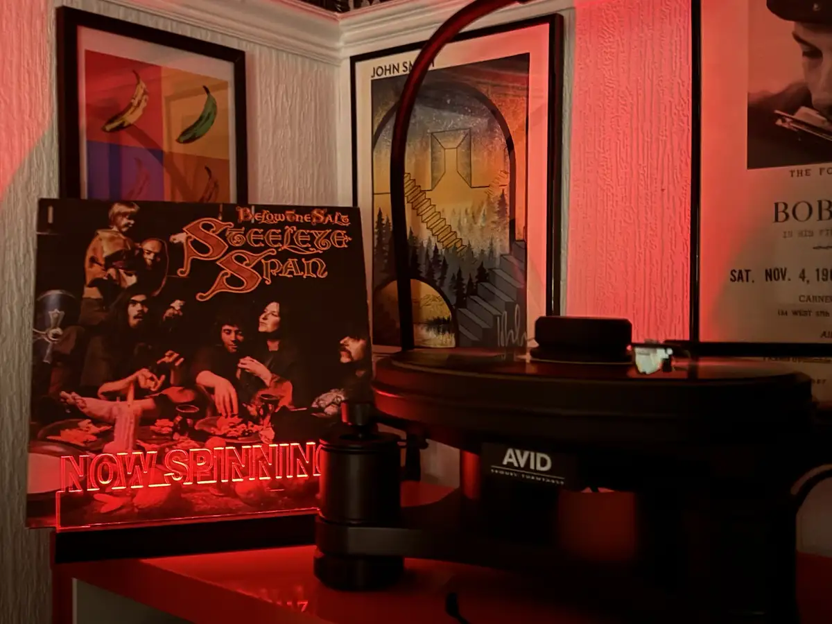 A vinyl record player with a glowing red 'NOW SPINNING' sign sits beside the 'Below the Salt' album by Steeleye Span. The…