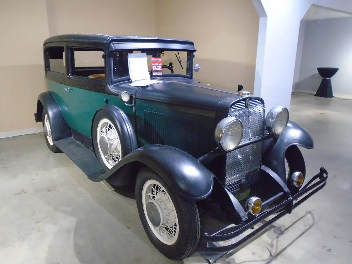 A vintage green and black automobile, likely from the 1930s, is displayed indoors on a concrete floor with beige walls. A…