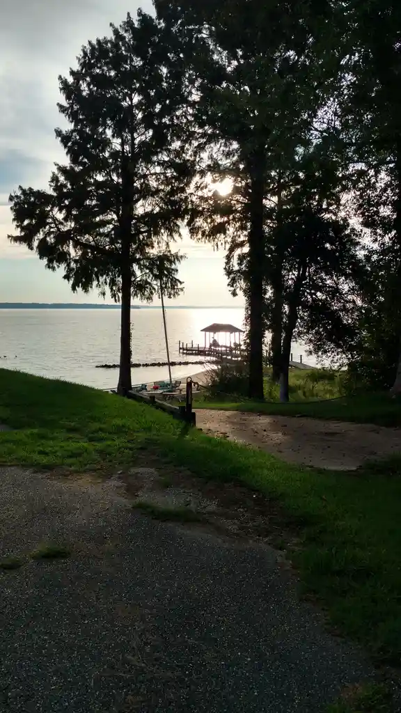 A serene lakeside scene at sunset, framed by tall trees, with a dock and gazebo visible in the distance. A gravel path leads…