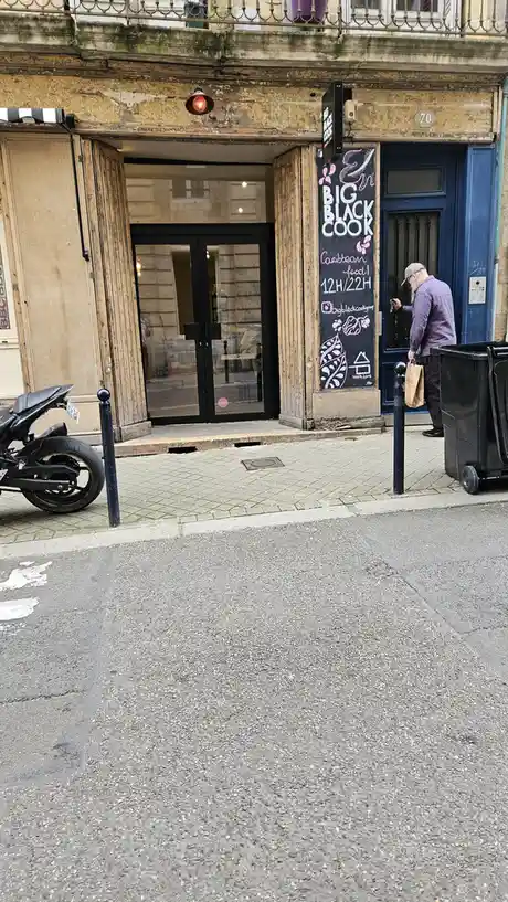 A man in a plaid shirt and cap stands at the blue door of a restaurant named 'Big Black Cook', which advertises Caribbean…