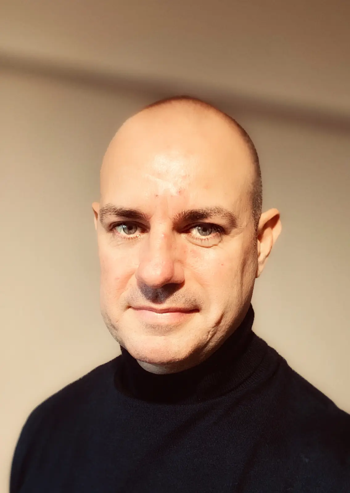 A close-up portrait of a bald man with light-colored eyes and a slight smile, wearing a dark turtleneck sweater against a…
