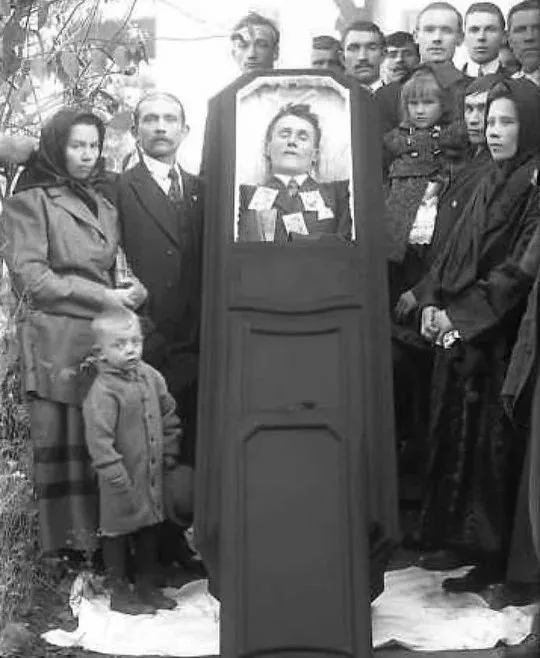 A black-and-white photo shows a group of people, including children, standing solemnly around an open coffin. Inside the…
