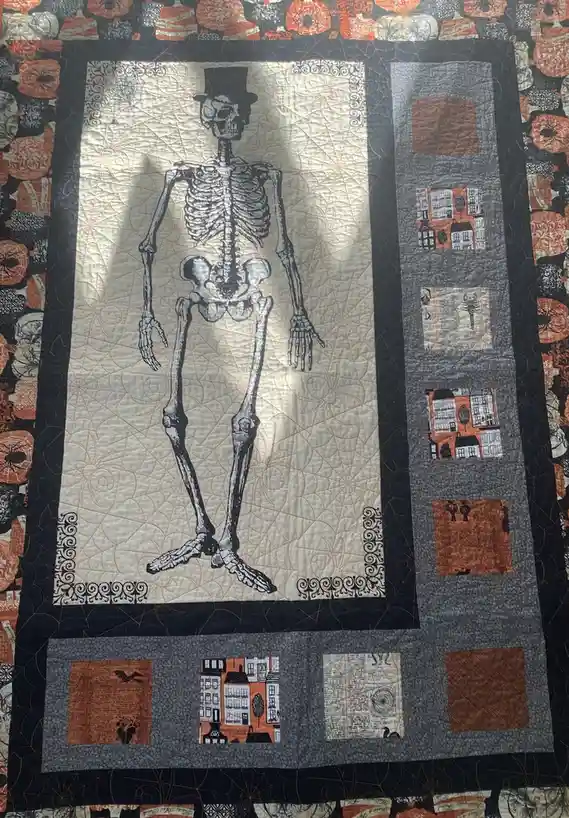 A Halloween-themed quilt features a large central panel with a full-body skeleton wearing a top hat, set against a light…