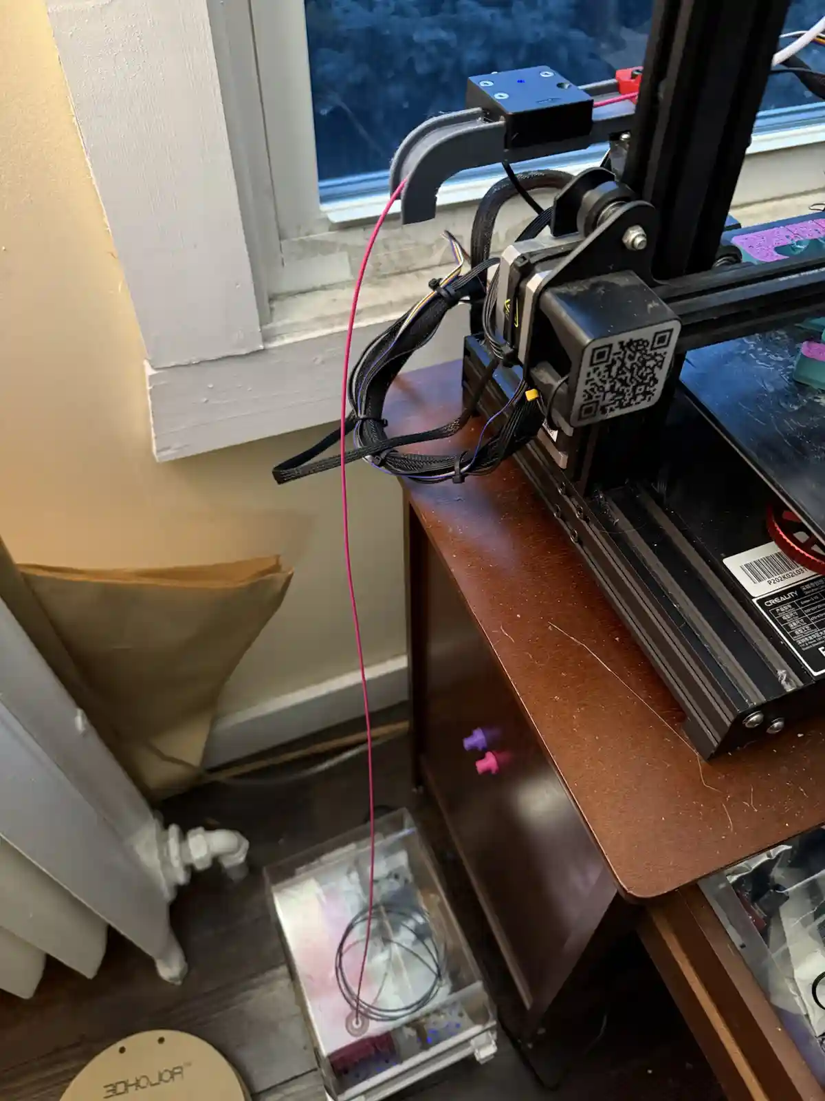 A 3d printer sits on a wooden desk near a window, with a pink filament cable dangling down to a clear plastic container…