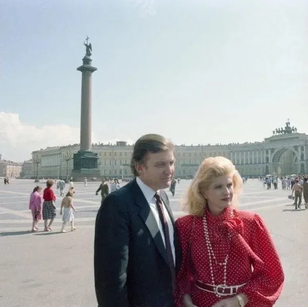 A vintage photo shows a man and woman standing together in a large public square with a tall column monument and grand…