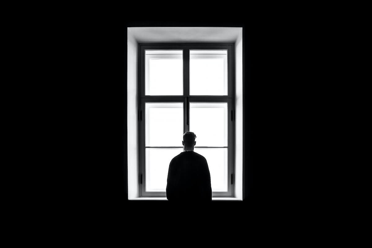 A black-and-white photo shows the silhouette of a person viewed from behind, standing in front of a bright, four-pane window…