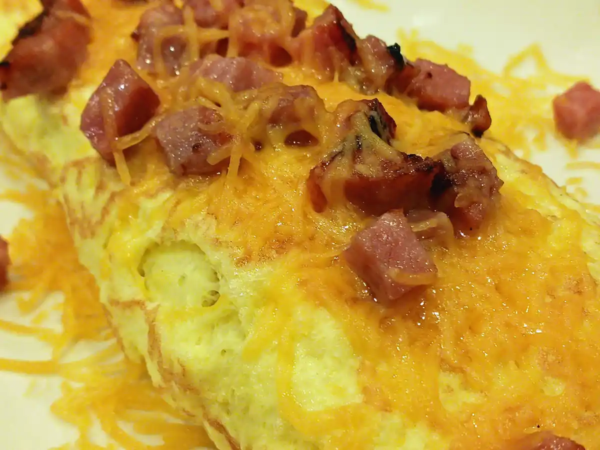 A close-up photo of a cheesy, ham-topped omelet on a white plate, with melted orange cheese and diced ham scattered across…