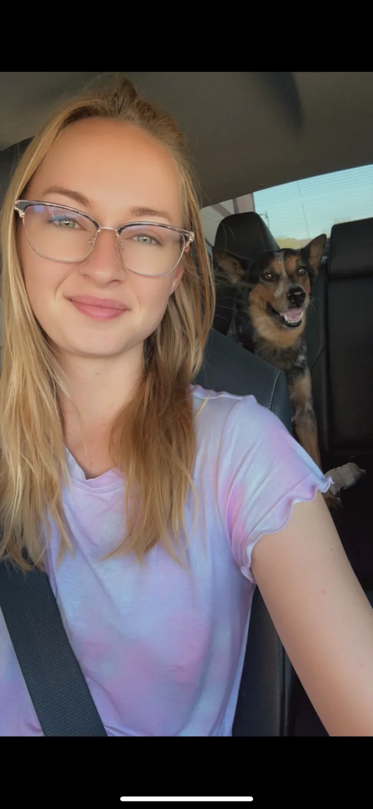 A selfie of a woman with blonde hair and glasses sitting in a car, smiling at the camera while wearing a seatbelt. Behind…