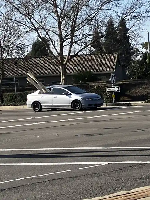 A white sedan is driving on a street with a 'ONE WAY' sign visible, but the car is loaded with long wooden planks sticking…