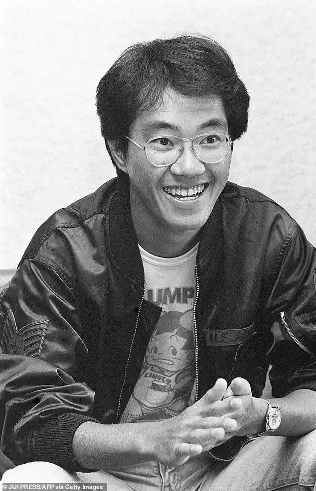 A black-and-white photo of a smiling man with glasses wearing a bomber jacket over a t-shirt with a cartoon character. The…
